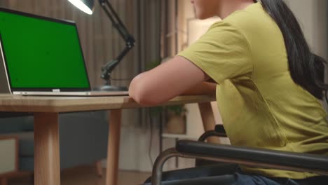 asian kid girl sitting in a wheelchair and raise hand before asking a question while learning online on laptop computer with mock up green screen at home