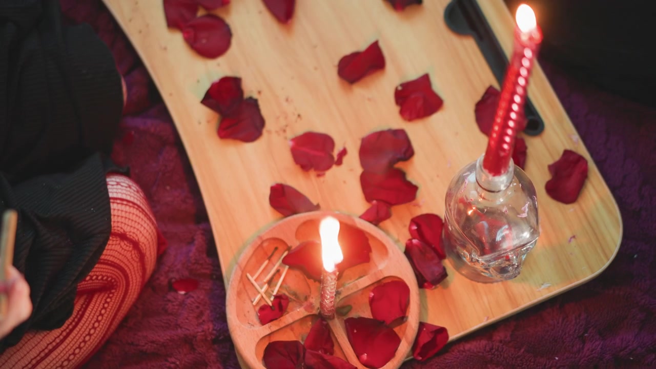 Bandeja de madera con pétalos de rosa esparcidos, vela roja encendida en botella de cristal, mano sosteniendo un fósforo, pétalos rodeando un pequeño cuenco, practicante preparando una lectura de amor en un ambiente cálido e íntimo de noche.
