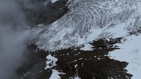 Drone-tilt-down-of-glacier-with-a-mix-of-ice-patterns-and-dark-rocks,-framed-by-swirling-clouds