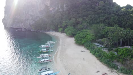 aerial view helicopter island tour c el nido - palawan : multiple banca tour boats anchored in turquoise water at white sand beach against tropical beach hut. light glare on camera, glitter on sea