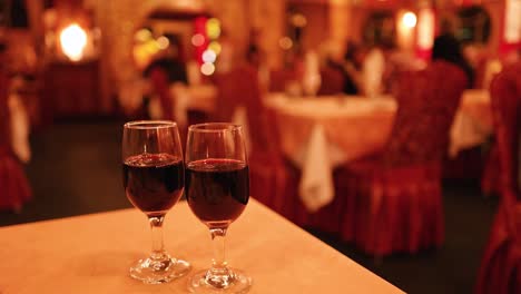 Two glasses of red wine rest on a table in a warmly lit, elegant restaurant with blurred diners and ornate decor in the background