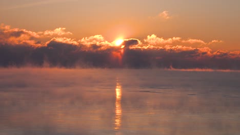 bright yellow red and orange sunrise through dark clouds over a very cold lake as steam rises in the middle of winter