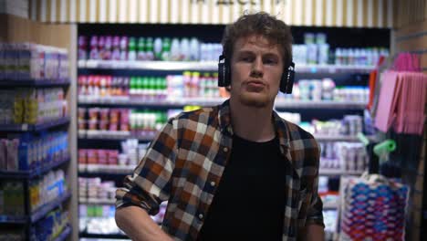 At-the-Supermarket:-Stylish-caucasian-guy-with-headphones-walks-through-goods-section-of-the-store-and-listening-to-the-music,-wearing-plaid-shirt.-Front-view.-Slow-Motion