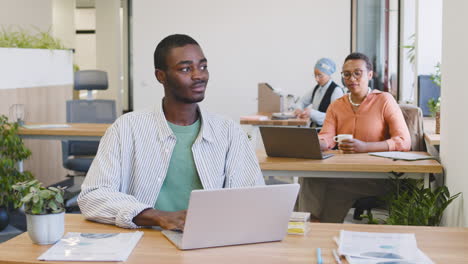 Junger-Lächelnder-Arbeiter,-Der-Mit-Einem-Laptop-Arbeitet,-Der-An-Seinem-Schreibtisch-Sitzt