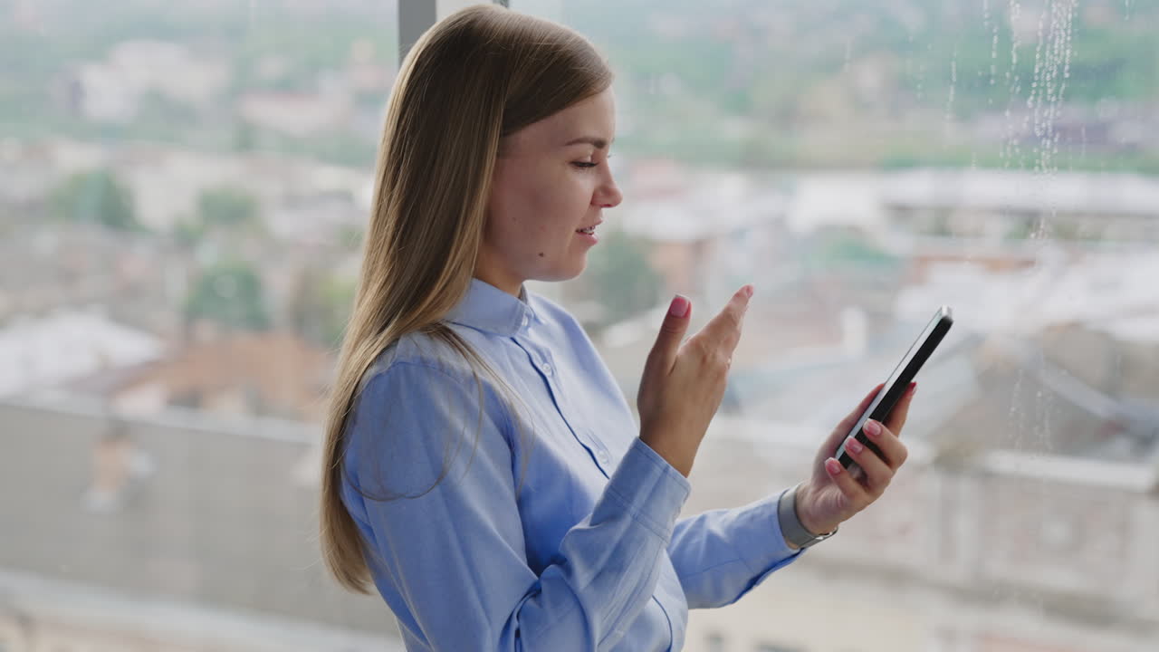 Beautiful blonde woman stands near window looking at her phone. Smiling woman having video chat on her smartphone. Panoramic window at backdrop.