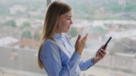 beautiful blonde woman stands near window looking at her phone. smiling woman having video chat on her smartphone. panoramic window at backdrop.