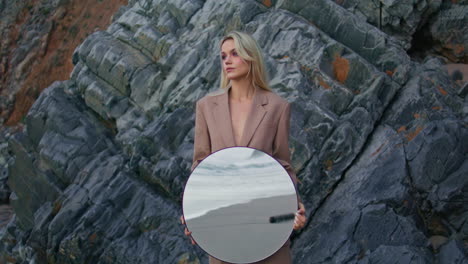 una chica de la orilla del mar posando rocas. una mujer atractiva y seria sosteniendo un espejo redondo.