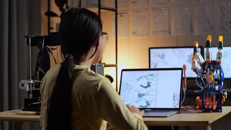 back view of teen asian girl with 3d printing having a backache while designing a cyborg hand on a laptop at home