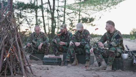 hombres militares con uniformes moro relajándose después de un turno de la tarde en una base en el campo, sentados en cajas afuera y preparando una fogata, recogiendo salchichas en palos, consumiendo alcohol