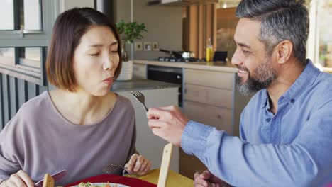 happy diverse couple sitting at table in dining room, eating dinner