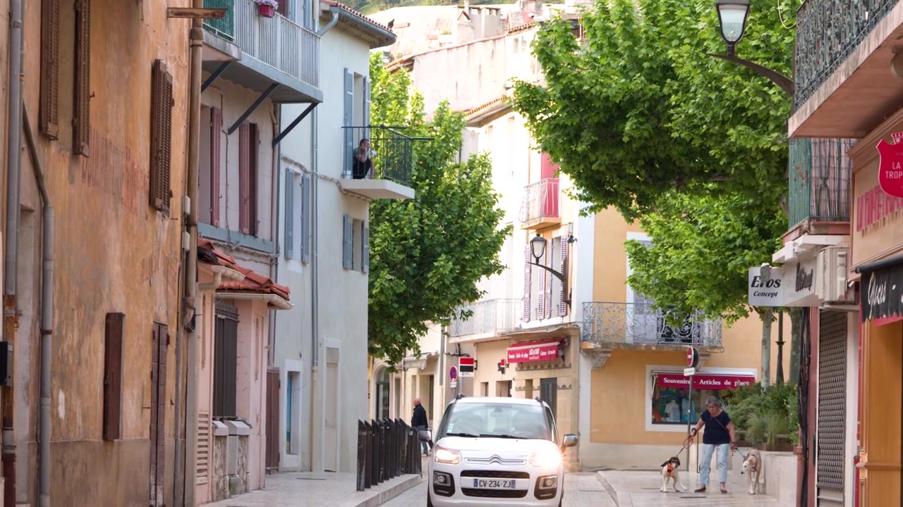 A white hatchback drives through a narrow Mediterranean alleyway lined with traditional French buildings.
