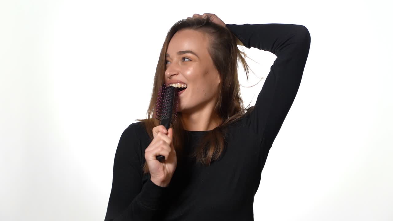 Smiling lady in black jumper and with long hair is holding a hairbrush in the studio on a white background.