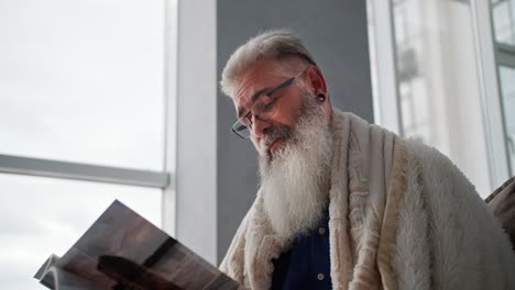 un hombre mayor con cabello gris y una barba exuberante y bien cuidada está envuelto en una manta blanca y leyendo una revista mientras está sentado en el sofá de un apartamento moderno