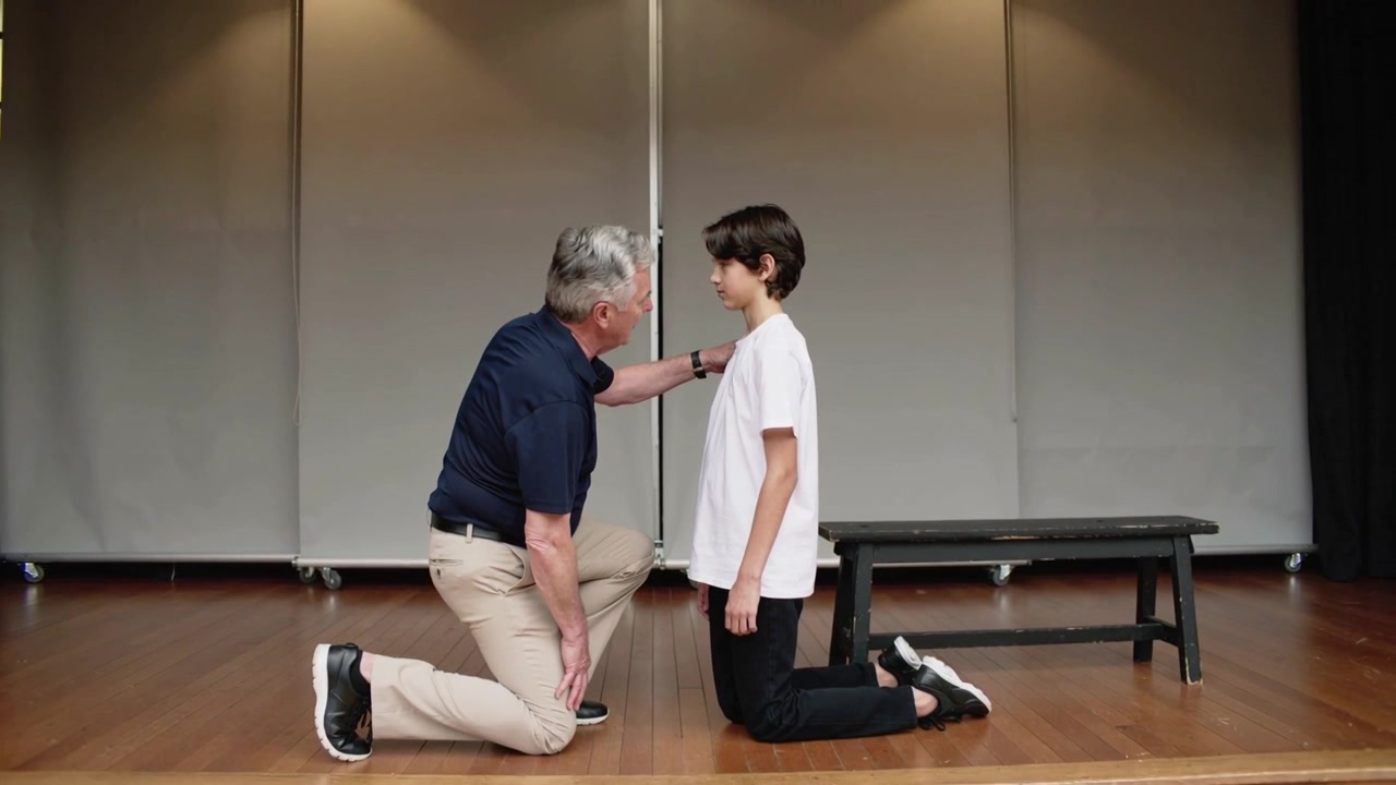 Walking left coach navy polo placing hand on teen, kneeling aligning heights guiding, bench stage