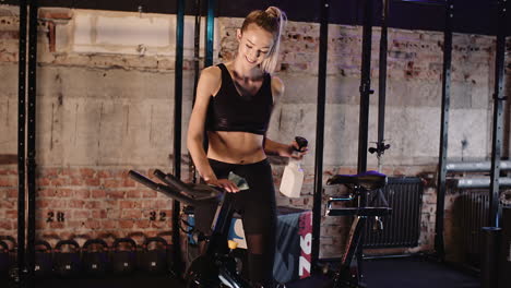 Smiling-Confident-Female-Instructor-Cleaning-Handle-Of-Stationary-Bike-At-Fitness-Club