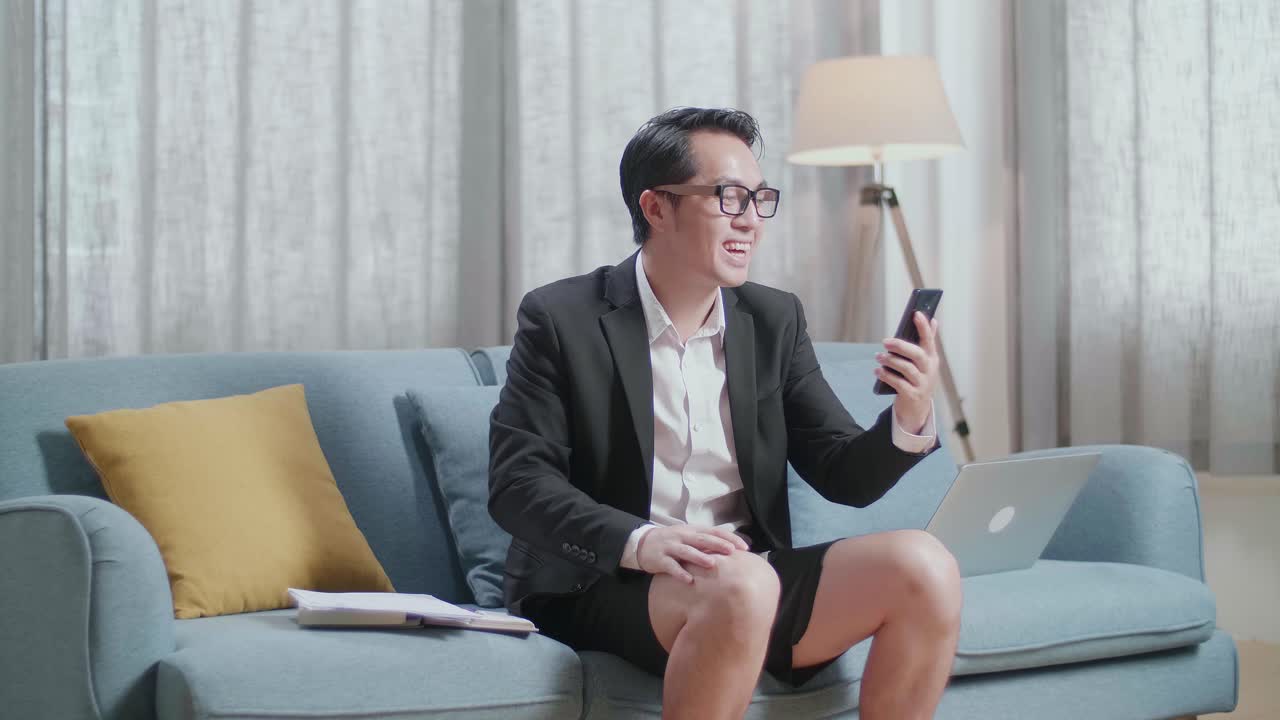 자켓과 쇼트를 입은 아시아 사업가 가 집에서 노트북으로 일하면서 스마트폰으로 비디오 통화를 하고 있다