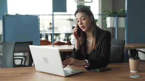 una mujer encantadora está sentada en la oficina, trabajando en su portátil y hablando por teléfono