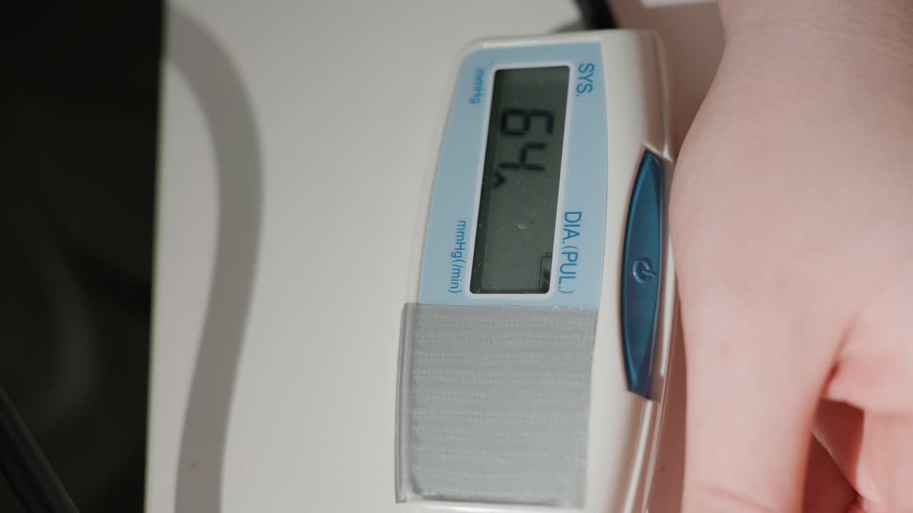Pulsossimetro da dito su mano bianca. L'illuminazione clinica costante rivela letture digitali che cambiano tra i fotogrammi mentre l'infermiera a domicilio monitora la saturazione di ossigeno e il polso, un morbido cinturino in tessuto lo fissa.