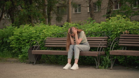 woman sits on wooden bench, holding head with both hands in somber and reflective posture, her long hair flows down, surrounded by lush greenery and trees, with building in background