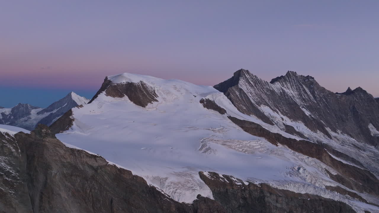 A breathtaking drone shot capturing the Alpuhbel, Täschhorn, and Dom peaks moments before sunrise, bathed in soft pink skies and glowing alpine light over the majestic Swiss Alps