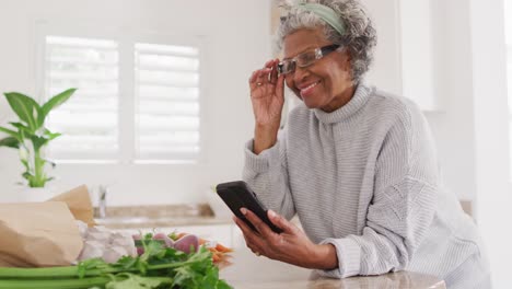Happy-senior-african-american-woman-cooking-in-kitchen,-using-smartphone