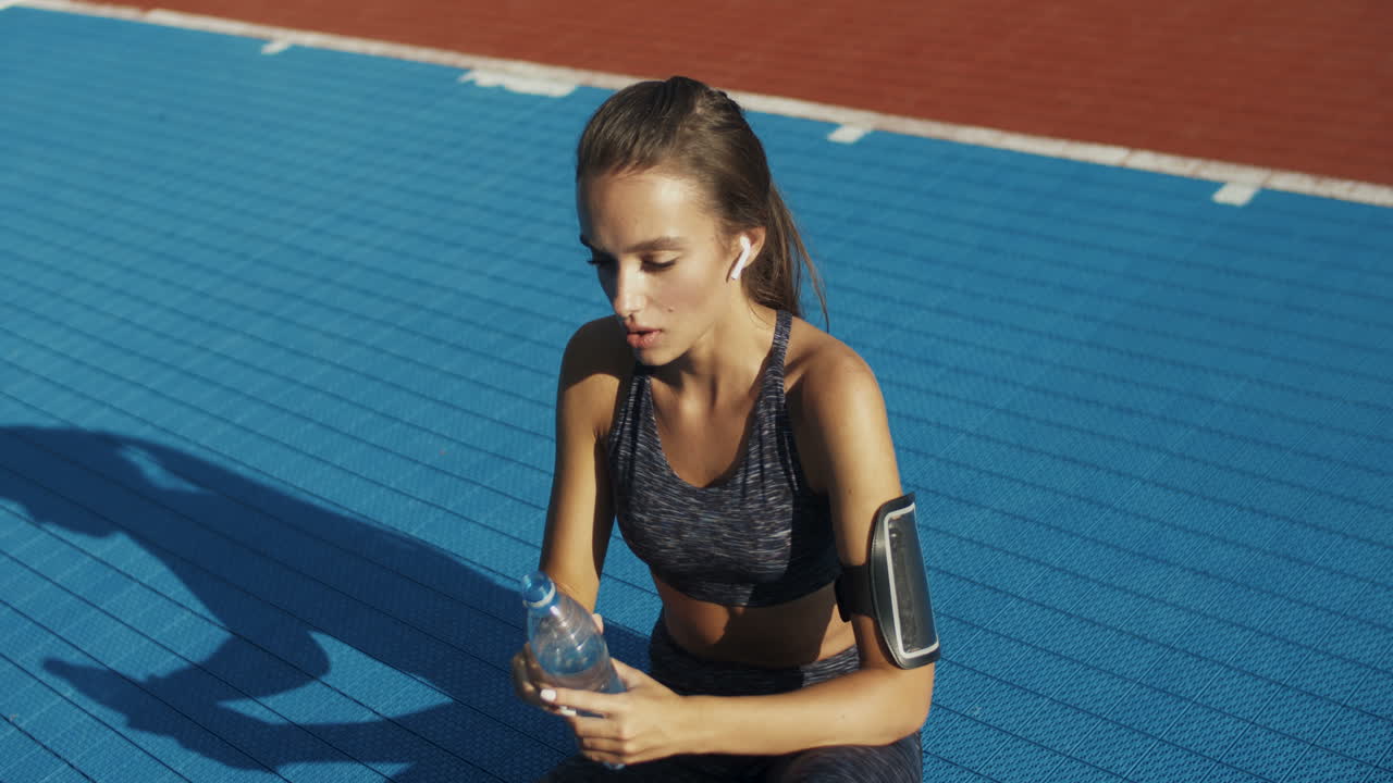 mujer fitness cansada sentada en la cancha deportiva con una botella de agua fría, limpiando el sudor de la frente y descansando después del entrenamiento