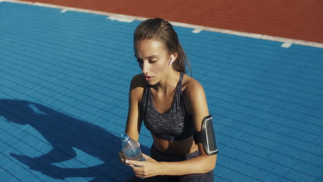 tired fitness woman sitting at sport court with bottle of cold water, wiping sweat from forehead and resting after workout