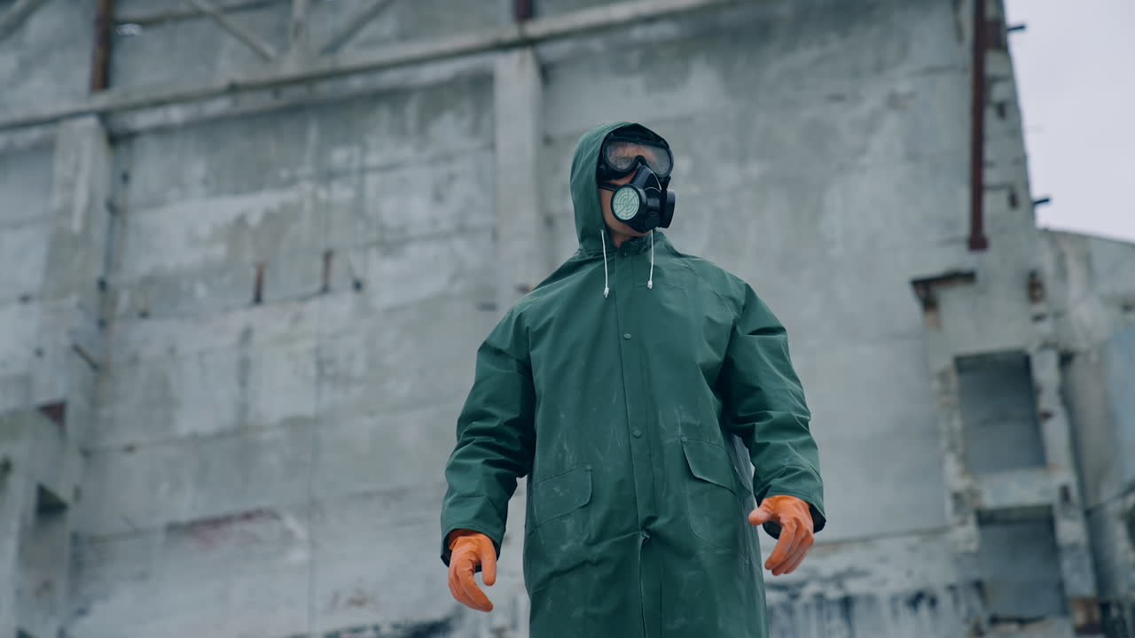 Human in protective costume and mask in infected place. Survivor man in a mask and a chemical protection suit showing on the camera with a finger.