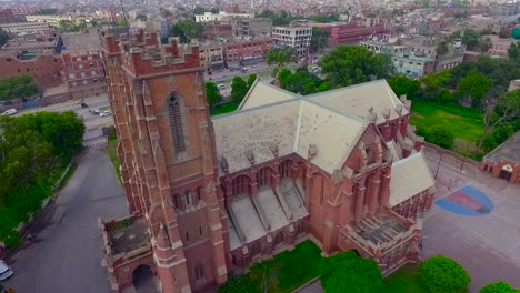 Vista-Aérea-De-Una-Hermosa-Iglesia-Antigua-En-La-Ciudad,-El-Tráfico-Se-Mueve-En-El-Otro-Lado-De-La-Calle-De-La-Iglesia,-Hermosa-Vegetación-Alrededor-De-La-Iglesia,-Una-Bandera-En-La-Parte-Superior-De-La-Iglesia