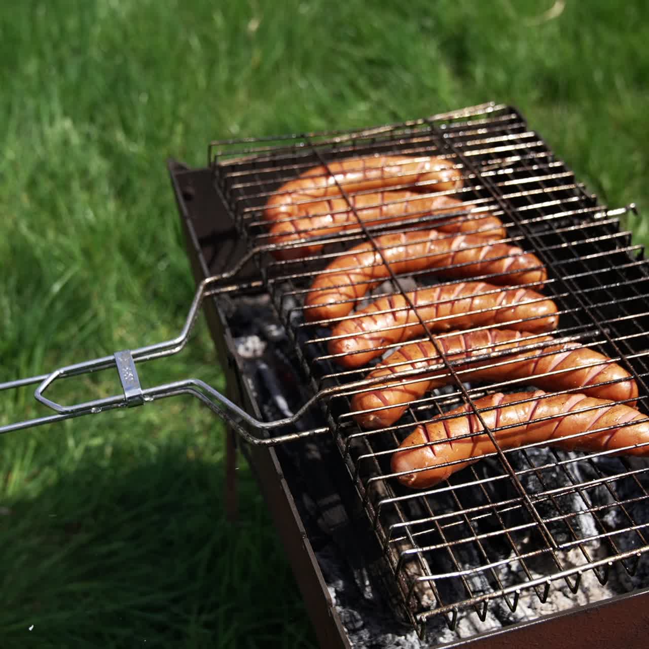 Grilling food for picnic. Delicious hot dog sausages frying in a grill grate in smoke on green nature background. Barbeque in summer