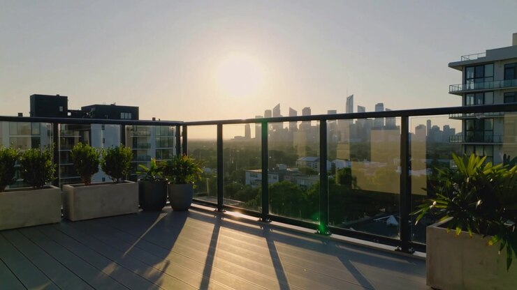 Sol em movimento sobre terraço na cobertura, guarda-corpo de vidro e vasos projetando sombras em deck composto, com reflexos.