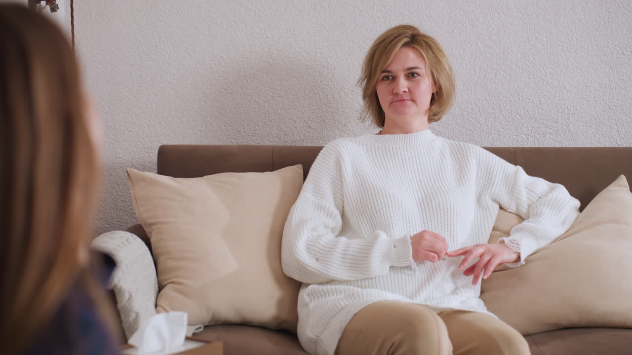 Participant seated on soft couch expresses point with hand gestures during thoughtful therapy exchange, facing professional listener in calm room with warm neutral tones and emotional clarity
