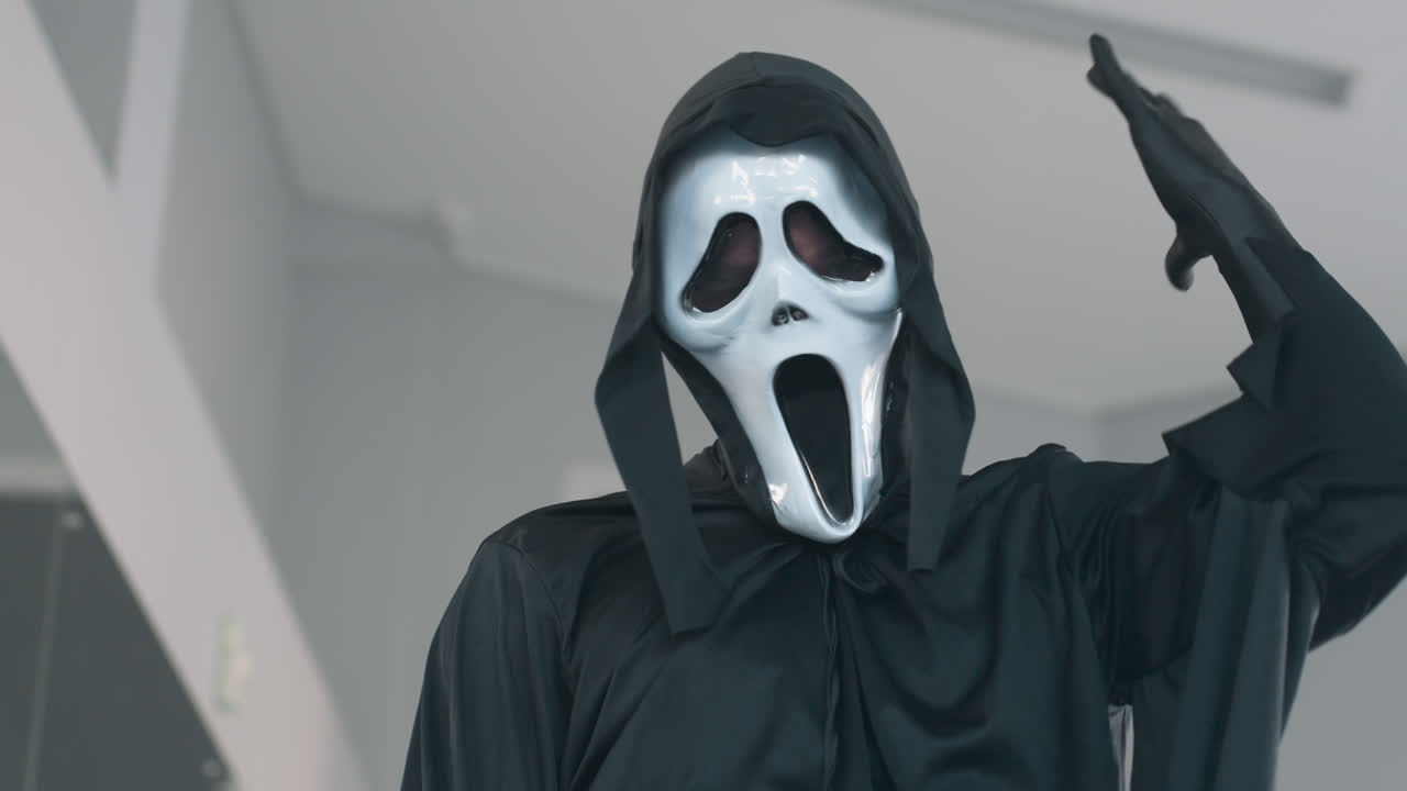 eerie masked figure in black hooded robe stands in dance studio mirror posing with dramatic gesture showcasing horror costume and glossy white ghost mask under soft window light