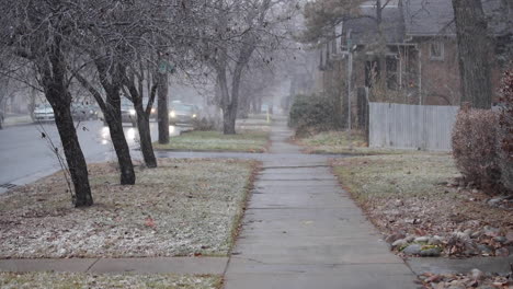 Nieve-Cayendo-En-La-Acera-De-Un-Barrio-Residencial-Mientras-Los-Coches-Pasan-A-Toda-Velocidad-Por-Una-Calle-Concurrida