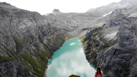 a reverse flyover above a hiker enjoying the view over lake limmernsee in glarus, switzerland, the turquoise colored water of which is surrounded by tall swiss alps peaks and cliffs