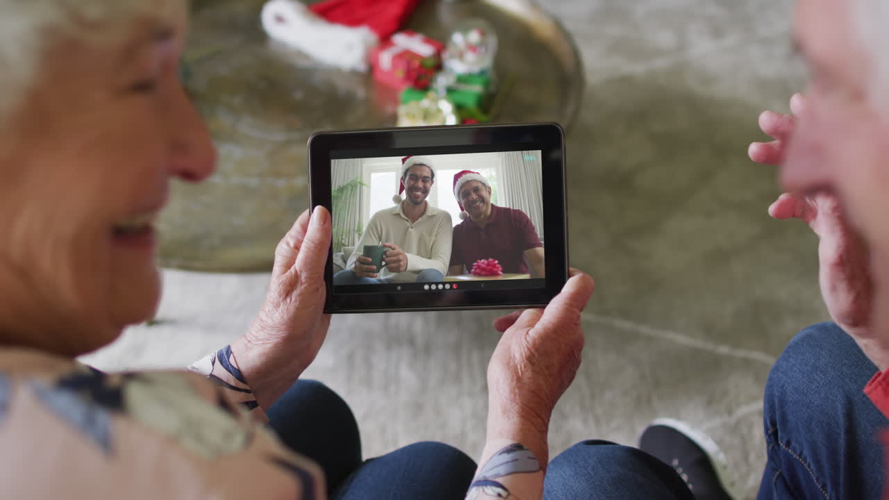 Coppia anziana caucasica sorridente che usa un tablet per una videochiamata di Natale con la famiglia sullo schermo