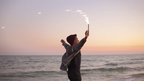 Young-handsome-african-guy-running-by-the-sea-during-sunset-and-holding-burning-sparkling-candle-in-his-hand.-Slow-Motion-shot