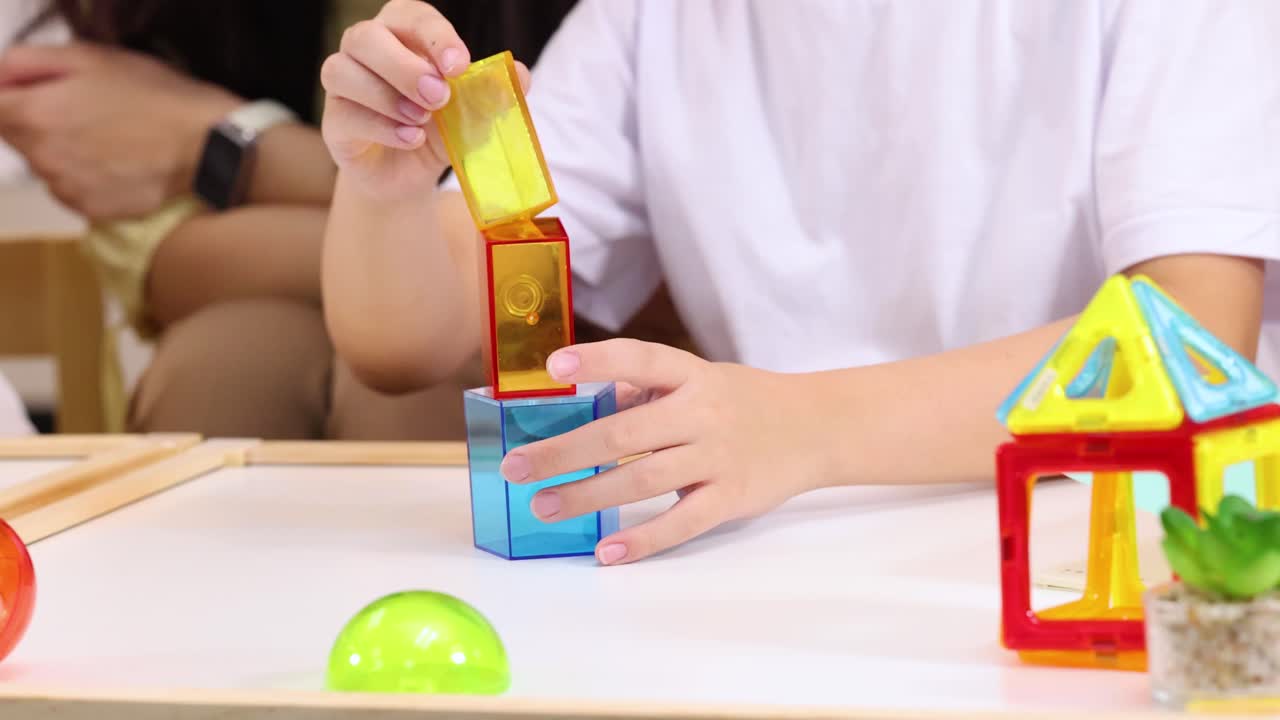 Child assembles translucent toy blocks at table, bright classroom lighting, teacher and classmates observing