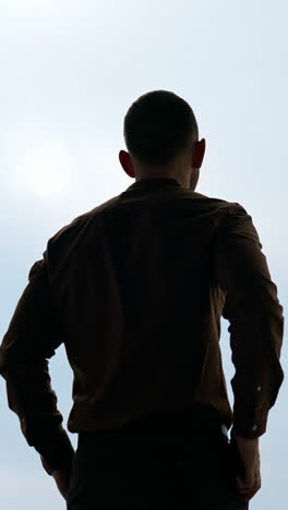young man on large window background. businessman with a phone standing in office and looking through the window in the evening. rear view. vertical video