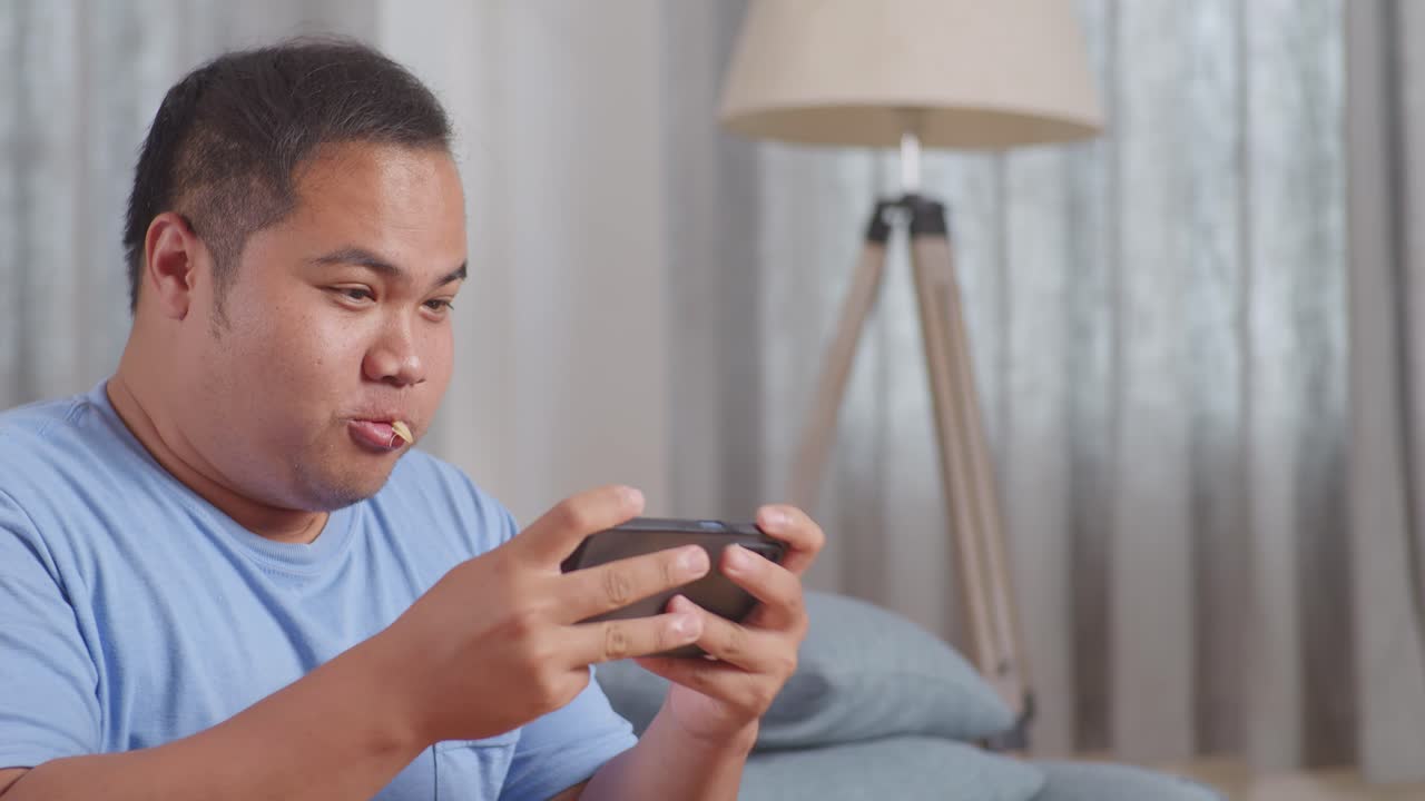 한 아시아 남자가 집에서 거실에서 소파에 긴 을 먹고 패스트푸드를 먹는 동안 스마트폰에서 우승 게임을 축하하는 장면