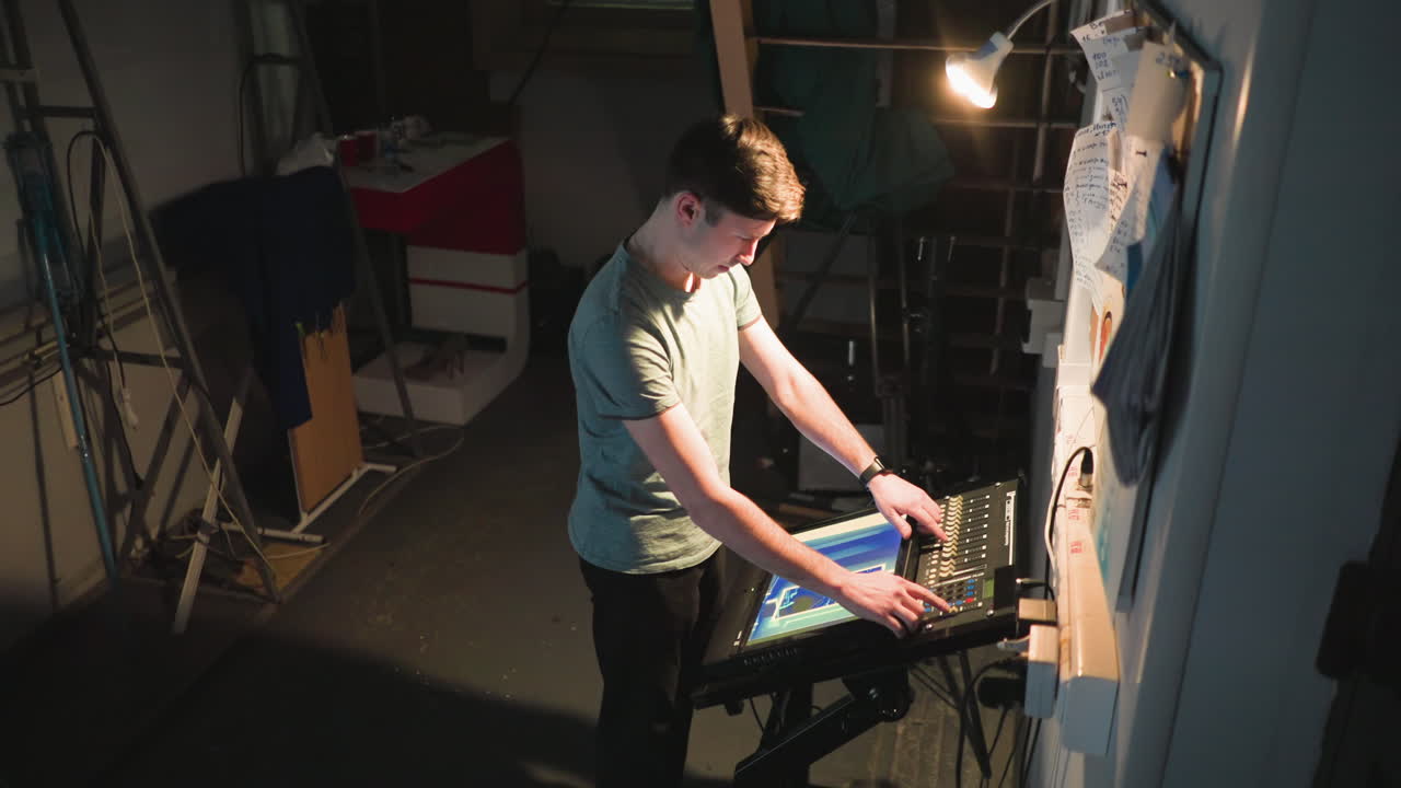 Young technician operates control panel under focused light in backstage area filled with cables, ladders, and bulletin board. Scene captures concentrated atmosphere of technical work in dim industrial space