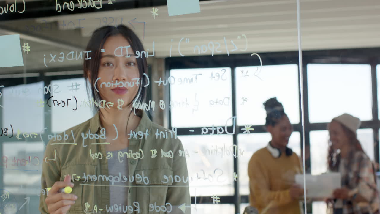 Woman writing code on glass board in modern office, collaborating with colleagues
