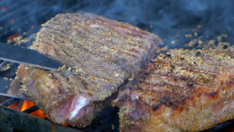 A-pair-of-meat-tongs-side-two-nearly-cooked-juicy-rib-eye-steaks-on-a-grill-while-flames-shoot-up-from-below