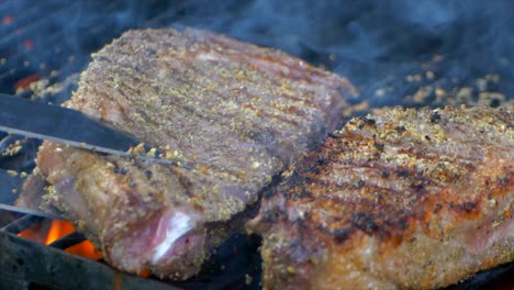un par de pinzas de carne al lado de dos jugosos filetes de costilla casi cocidos en una parrilla mientras las llamas se disparan desde abajo