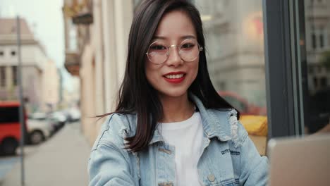 portrait of the beautiful young girl in stylish look and glasses typing on the laptop computer while sitting in the cafe summer terrace and then smiling to the camera