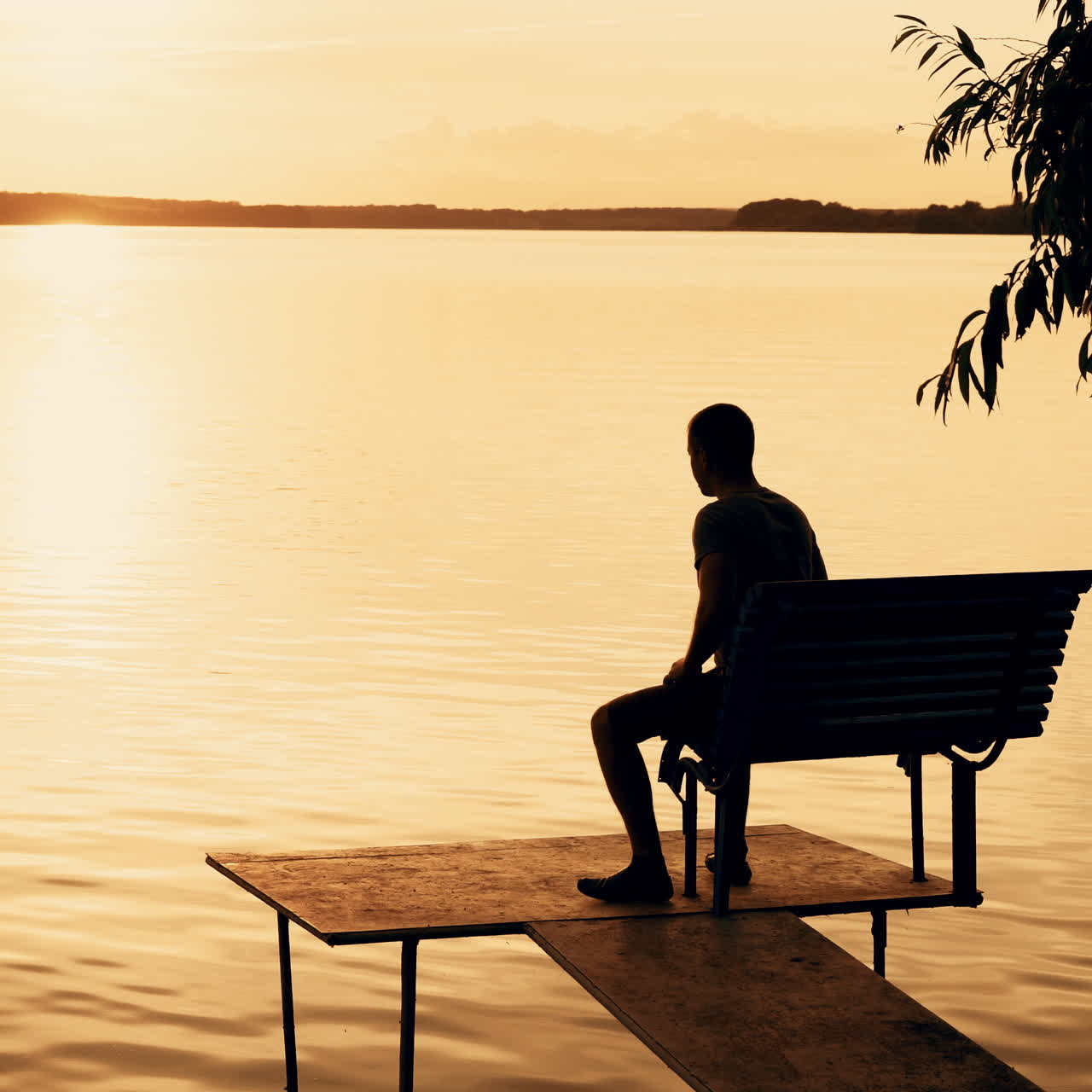 A man was standing on the bridge and than sat down on a bench to looking at the beauty of nature on the background of the river and the sunset on a summer evening.