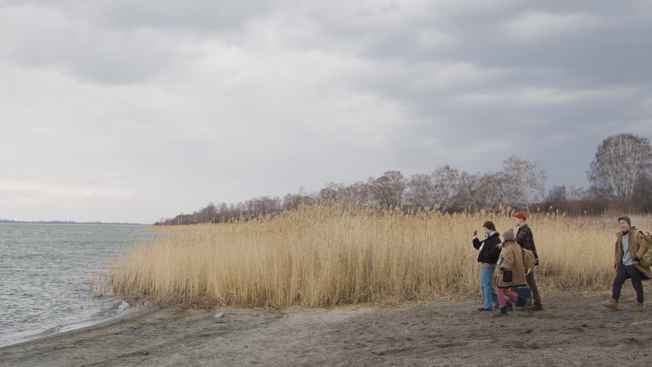 вид сбоку на группу мальчиков и девочек-подростков в зимней одежде, идущих по пшеничному полю недалеко от моря в пасмурный день