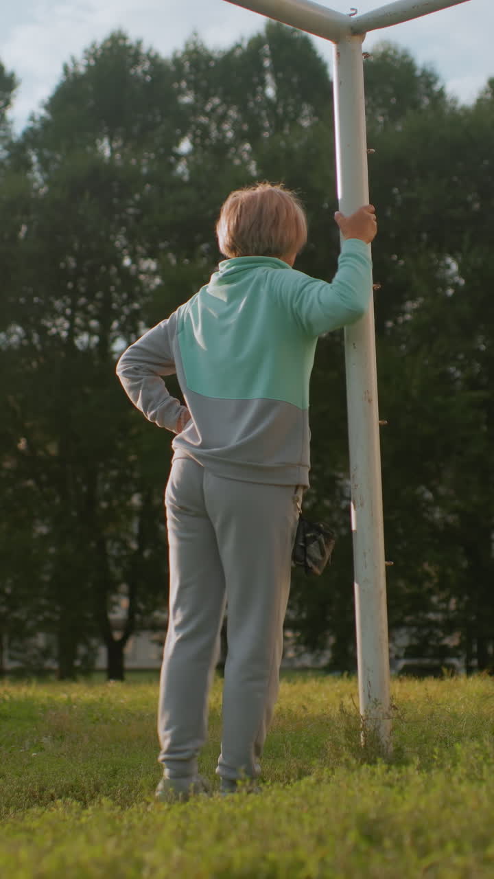 Anziana donna bianca che si allunga al palo della porta, solleva la gamba appoggiata al palo e si arrampica giocando in un parco giochi, luce del sole serale, mani che afferrano il palo, mettendo alla prova l'agilità e la resilienza con un cane fedele nelle vicinanze