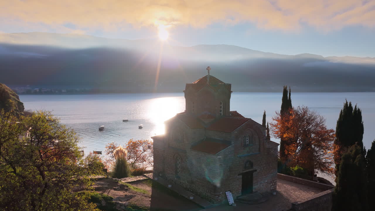 Aerial drone view of a sunrise scene behind St. John at Kaneo, with rays breaking through the clouds and illuminating the church's silhouette. North Macedonia