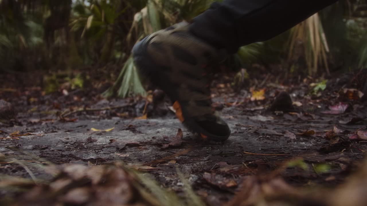 A close-up of a rugged hiking boot stepping on a muddy forest trail surrounded by fallen leaves and dense greenery. The scene evokes a sense of adventure, exploration, and connection with nature